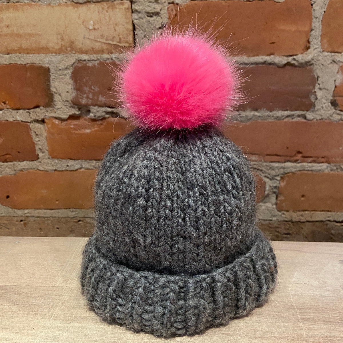 A dark gray chunky knit beanie with a fluffy hot pink pom-pom, displayed on a light wood surface against a rustic red-brown brick wall background.