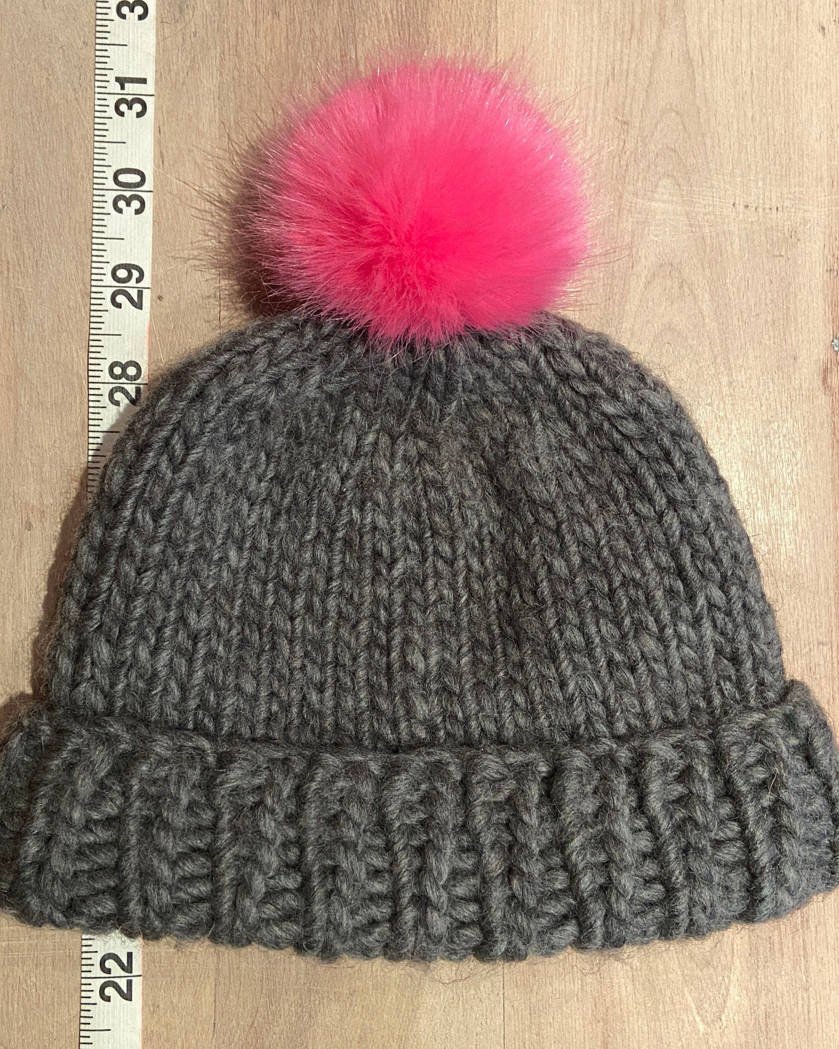 A gray chunky knit beanie with a bright pink fluffy pom-pom, laid flat on a light wood surface next to a measuring tape.
