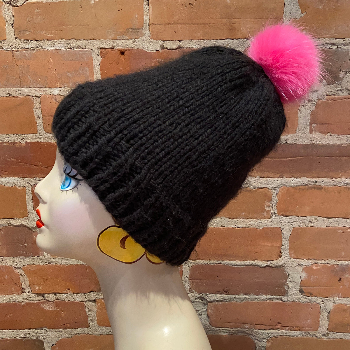 A black ribbed knit beanie with a bright pink faux-fur pom-pom, displayed on a mannequin head against a red-brown brick wall background.