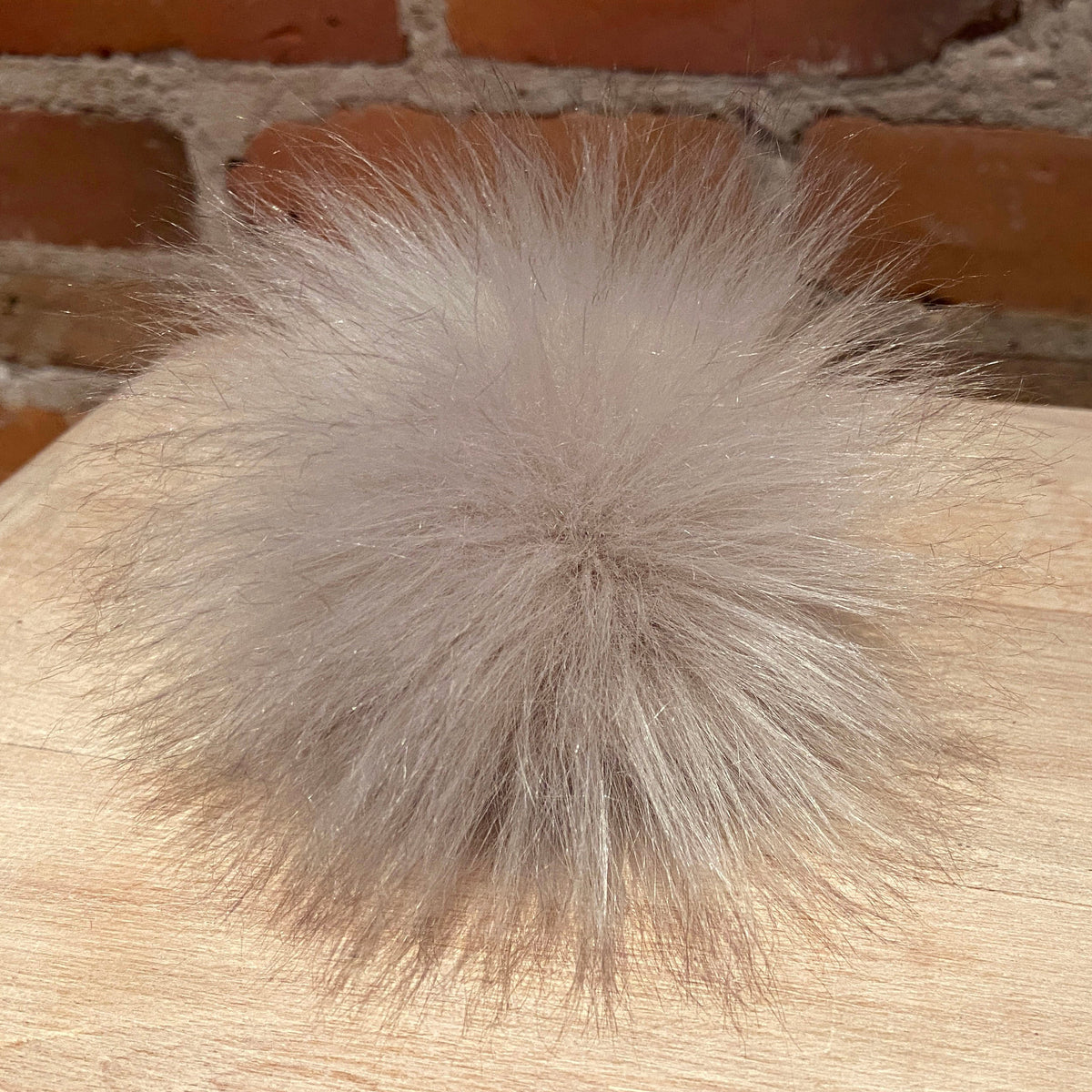 A fluffy faux fur pom-pom ball in a light taupe-gray color, resting on a light wood surface with a brick wall background.