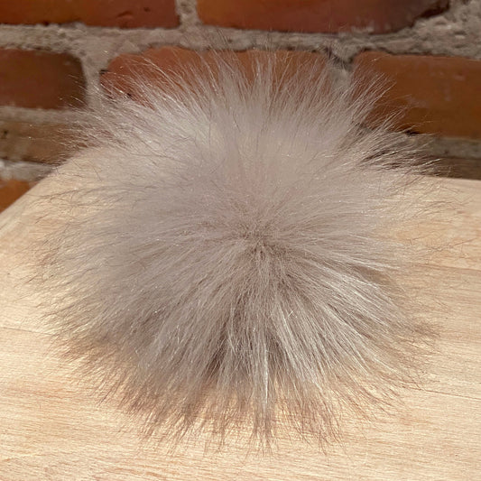A fluffy faux fur pom-pom ball in a light taupe-gray color, resting on a light wood surface with a brick wall background.
