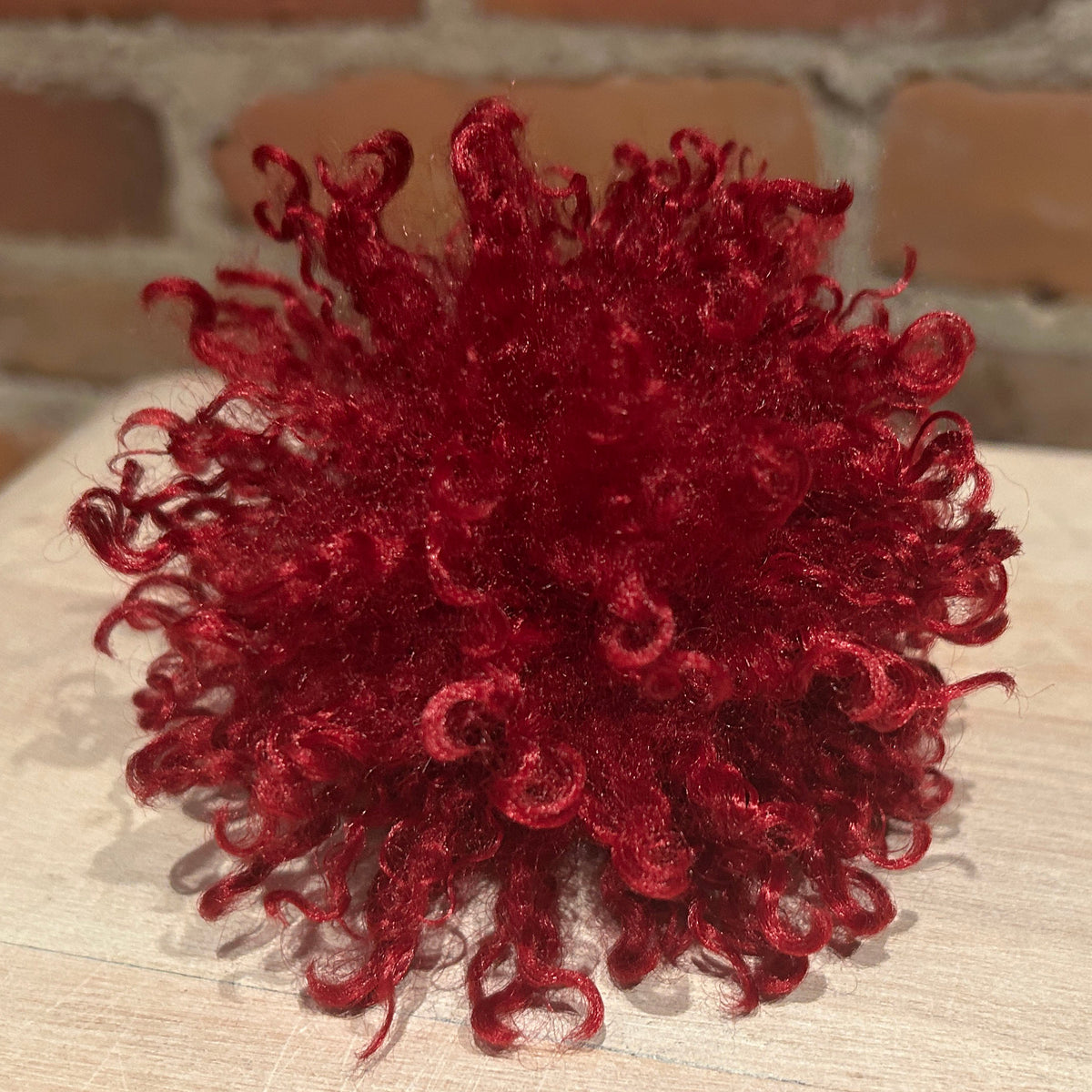 A deep red curly faux fur pom-pom toy or decorative accessory, displayed on a light wood surface with a brick wall background.