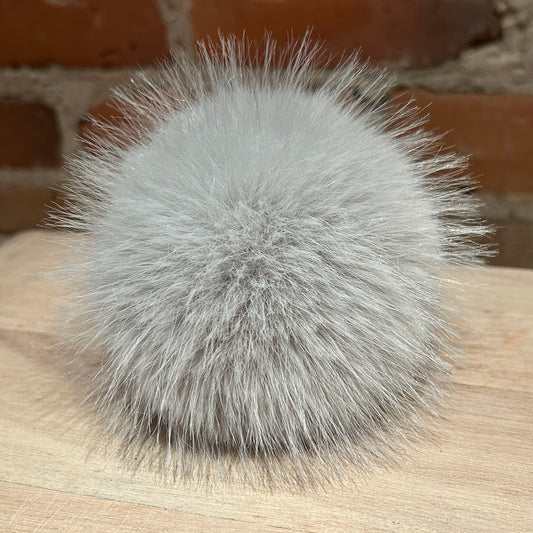 A fluffy light gray faux fur pom-pom accessory resting on a light wood surface with a blurred brick wall background.