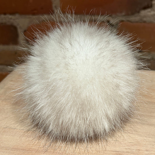 A fluffy white faux fur pom-pom ball resting on a light wood surface with a blurred brick wall background.