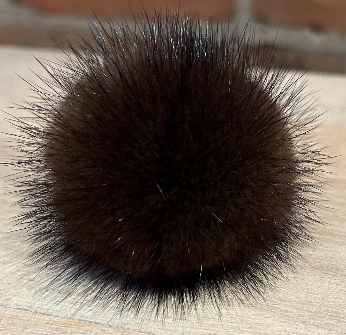 A close-up of a dark brown mink fur pom-pom accessory, likely a hair claw clip or decorative ball, resting on a light wood surface with a blurred brick wall background.