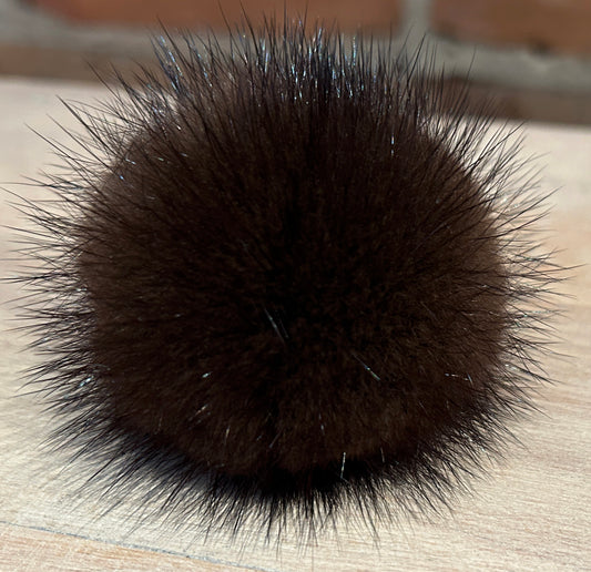 A close-up of a dark brown mink fur pom-pom accessory, likely a hair claw clip or decorative ball, resting on a light wood surface with a blurred brick wall background.