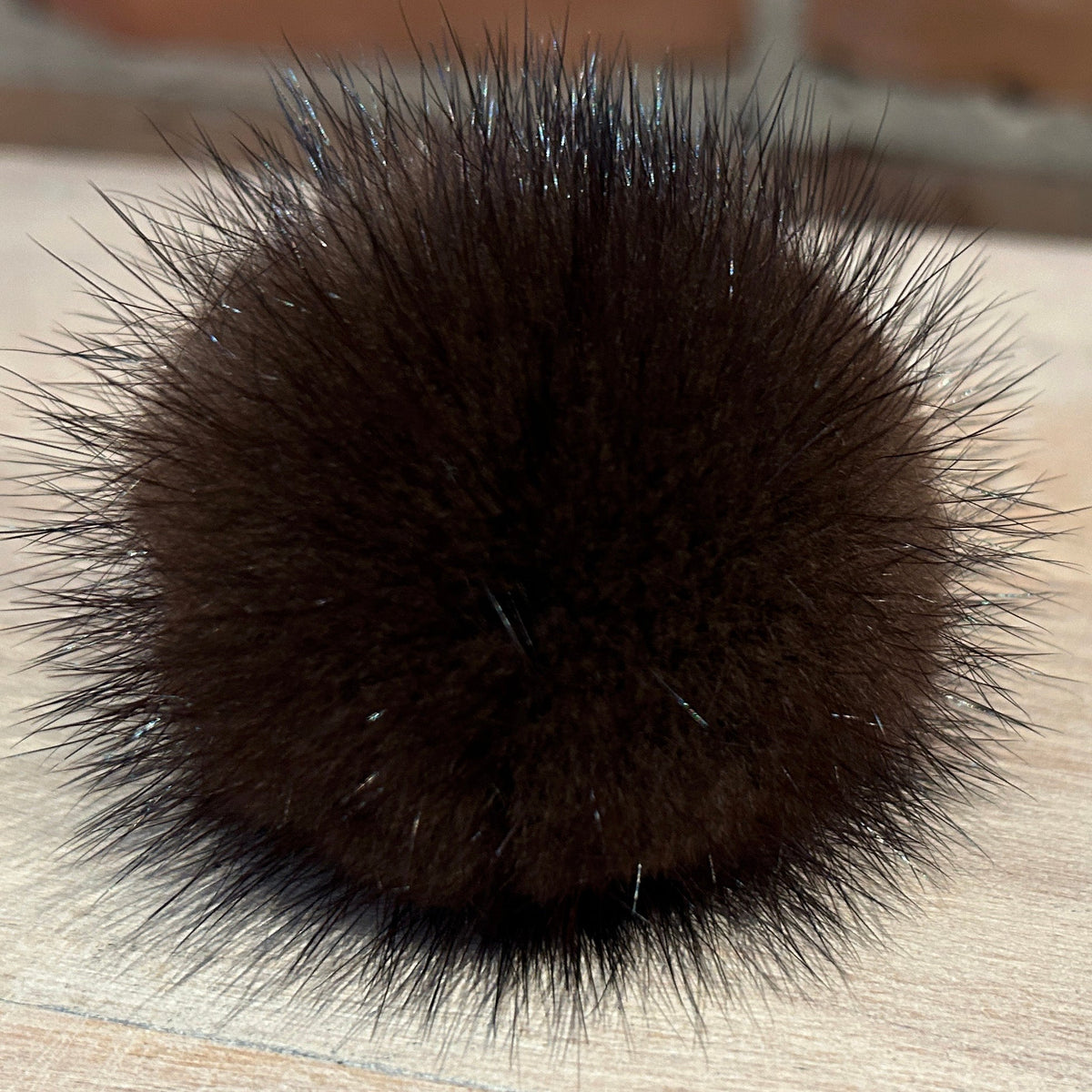 A close-up of a dark brown mink fur pom-pom accessory, likely a hair claw clip or decorative ball, resting on a light wood surface with a blurred brick wall background.