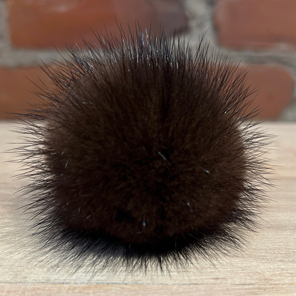 A dark brown mink fur pom-pom ball resting on a light wood surface with a blurred brick wall background.