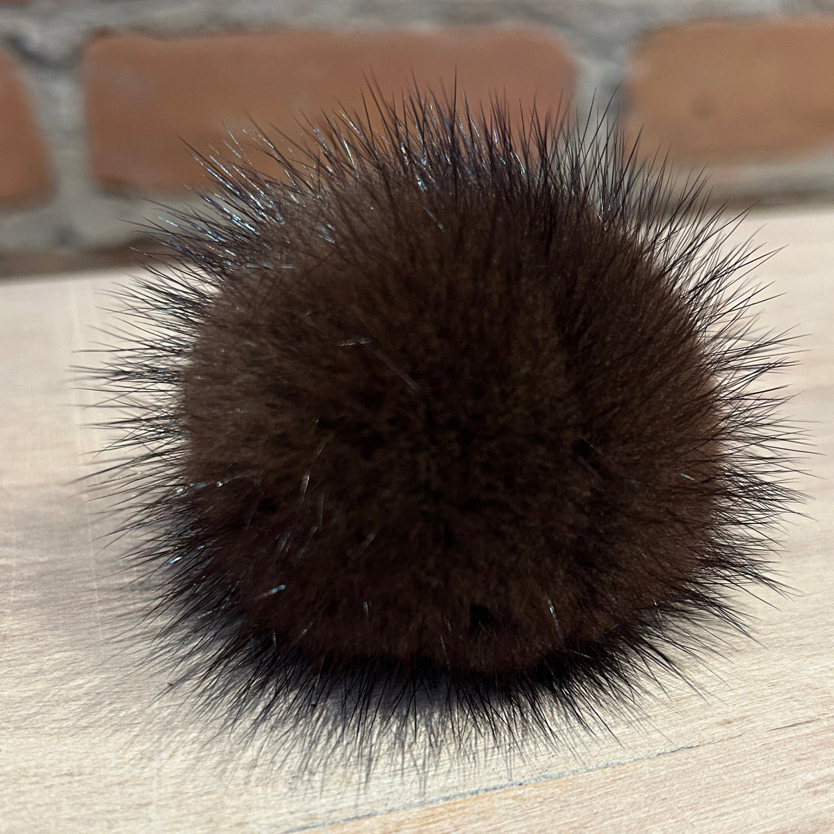 A dark brown mink fur pom-pom accessory with spiky, voluminous fibers, resting on a light-colored wooden surface with a blurred brick wall in the background.