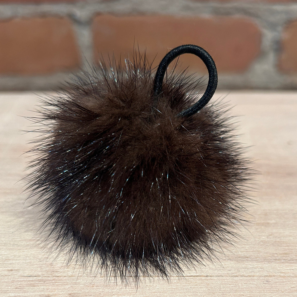 A dark brown fluffy mink fur pom-pom keychain with a black elastic loop, resting on a light wood surface with a blurred brick wall background.