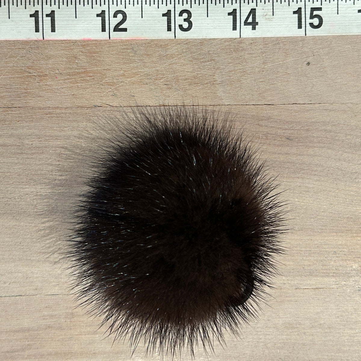 A dark brown fluffy pom-pom accessory placed on a light wood surface with a measuring ruler visible along the top edge for scale.