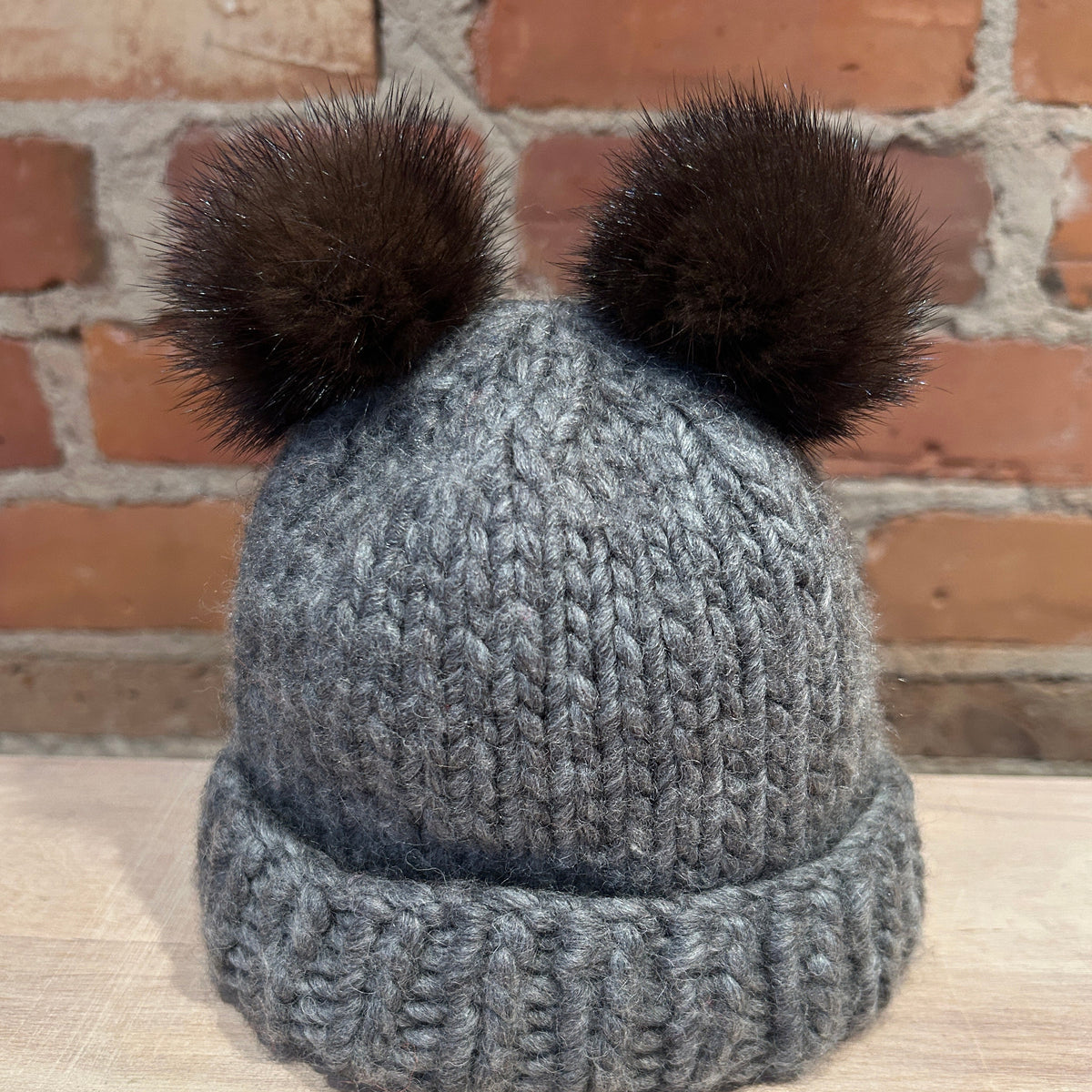 A gray chunky knit beanie hat with two dark brown mink fur pom-poms on top, displayed on a light wooden surface against a brick wall background.
