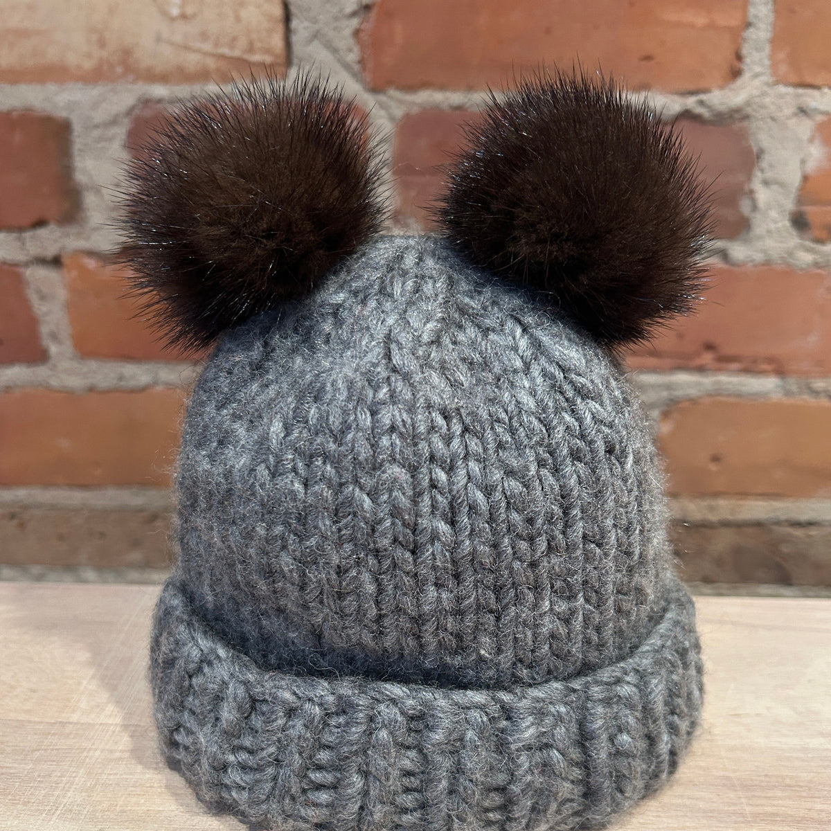 A gray chunky knit beanie hat with two dark brown mink fur pom-poms on top, displayed on a light wooden surface against a brick wall background.