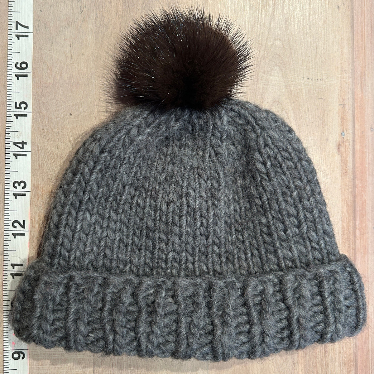 A gray chunky knit beanie with a dark brown mink fur pom-pom, laid flat on a light wood background next to a measuring tape.