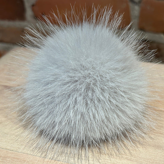 A fluffy light gray faux fur pom-pom accessory resting on a light wood surface with a blurred brick wall background.