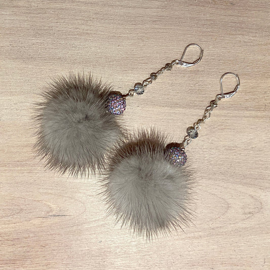A pair of dangle earrings featuring large, fluffy gray mink fur pom-poms, connected by silver-toned beads and a small iridescent purple rhinestone ball, hanging from silver fishhook ear wires on a light wood background.