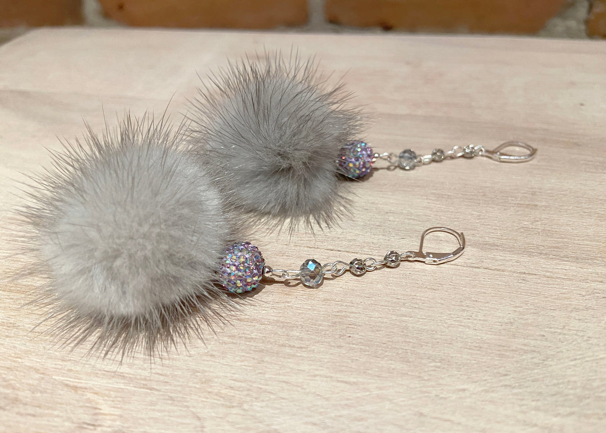 A pair of dangle earrings featuring large fluffy light gray mink fur pom-poms, connected by silver-toned chains with iridescent rhinestone beads, displayed on a light wood surface.