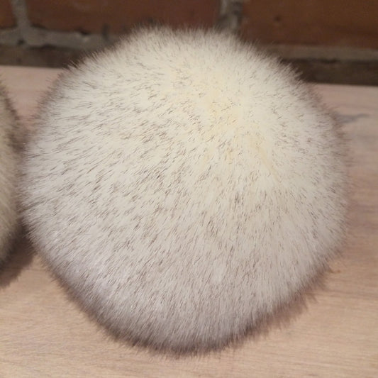 A round, fluffy faux fur or rabbit fur pad in a light cream-white color, resting on a light wood surface with a brick wall in the background.