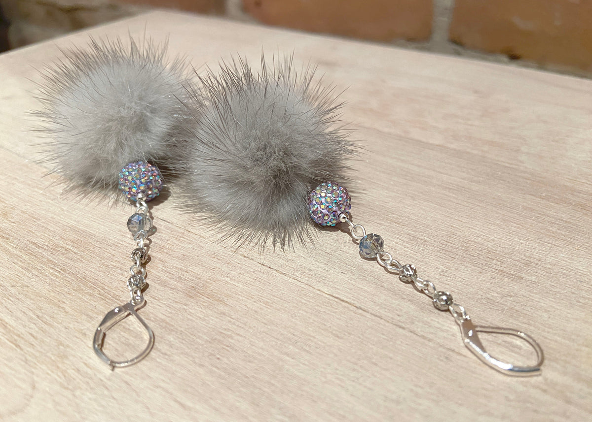 A pair of long dangle earrings featuring fluffy light gray mink fur balls, a small iridescent rhinestone bead, a clear faceted crystal, and silver-tone chain links ending in hook clasps, displayed on a light wood surface.