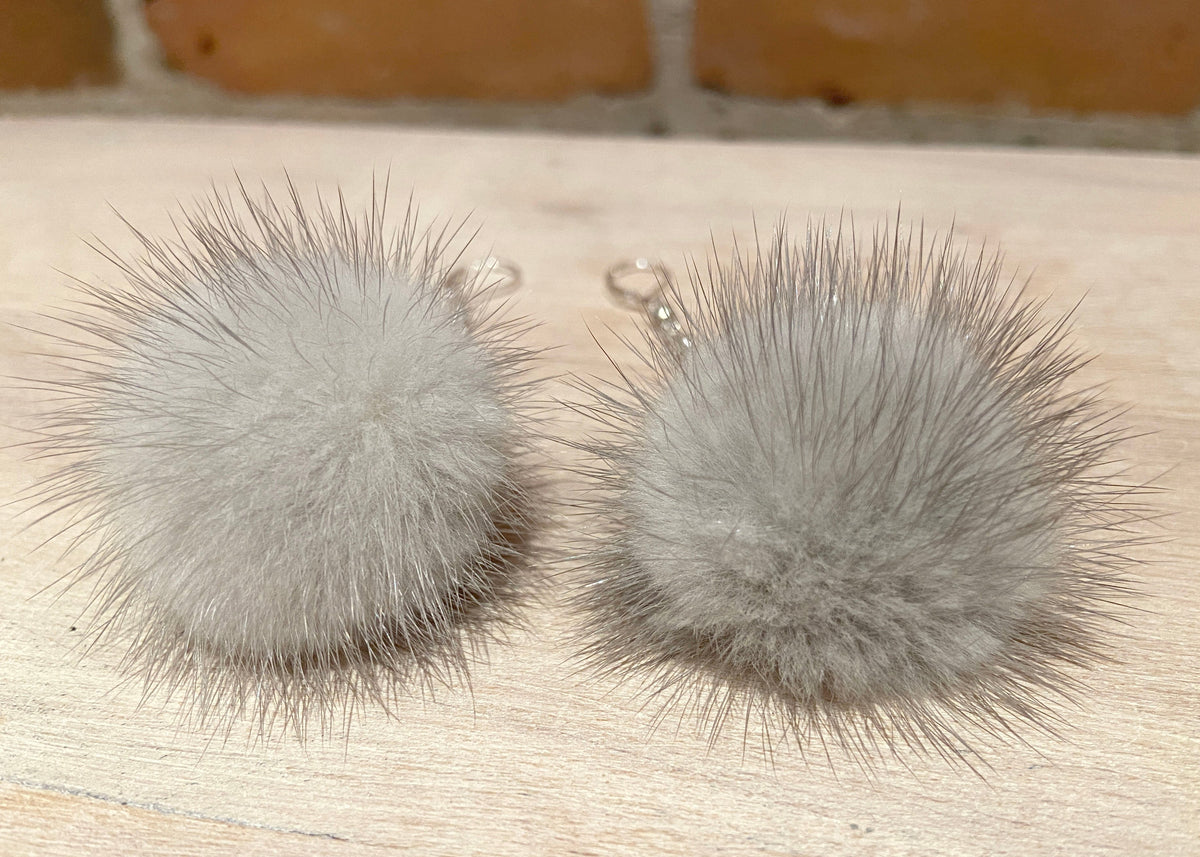 Two fluffy gray mink -fur pom-pom keychains with silver lobster clasps, resting on a light wood surface with a brick wall background.