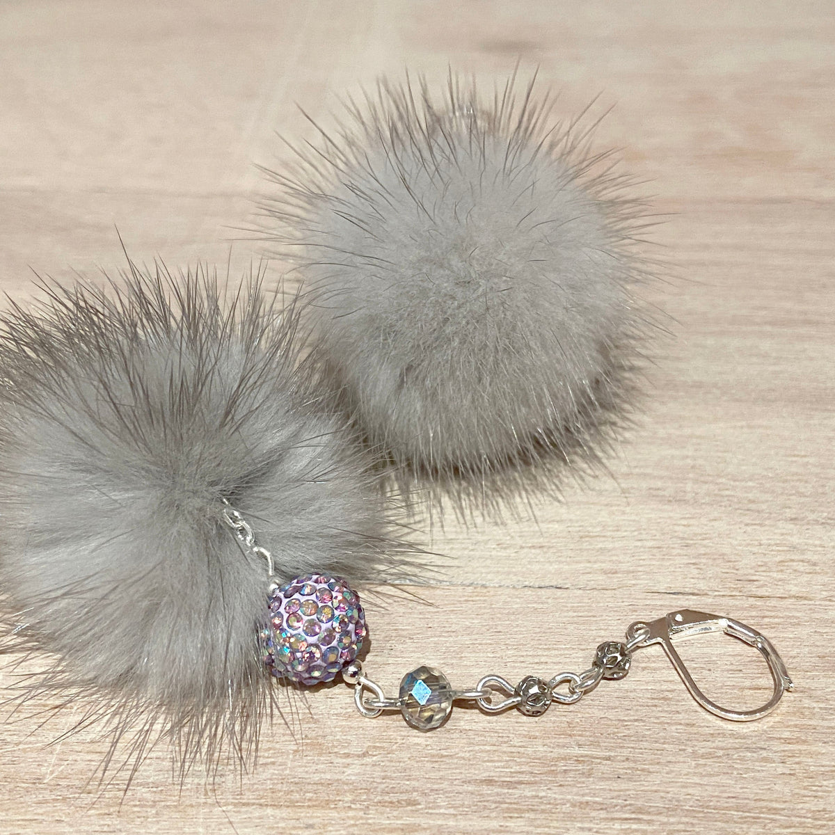 A pair of gray fmink aux fur pom-pom keychains with a silver-tone chain, a glittery multicolor bead, and a small iridescent bead on a light wood background.