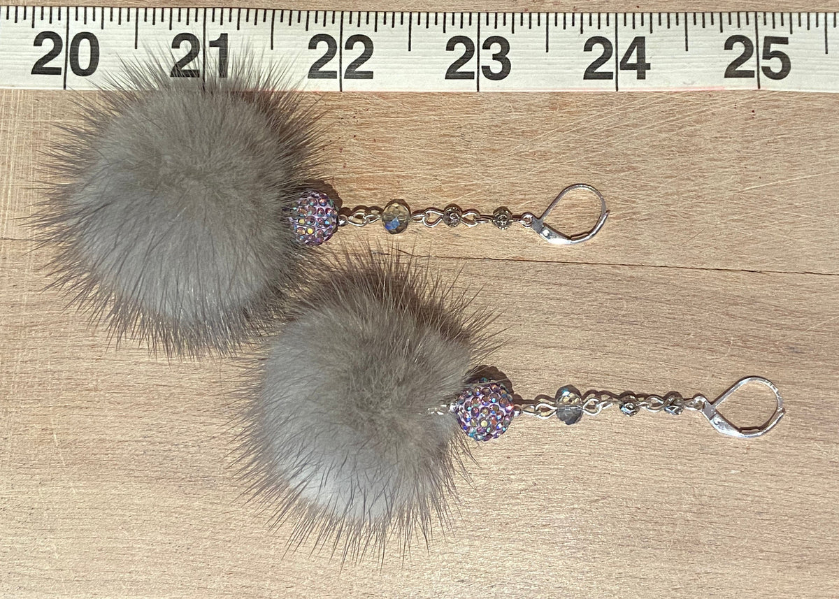 A pair of dangling earrings with large fluffy gray mink -fur pom-poms, silver-tone hooks, and small iridescent rhinestone beads, shown on a wooden surface next to a ruler for scale.
