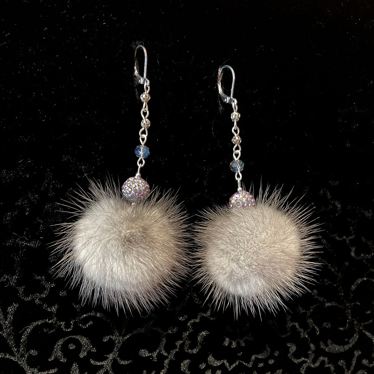A pair of dangle earrings featuring large, fluffy light beige mink fur pom-poms, connected by silver chains to silver fishhook ear wires. Each chain is accented with a small blue faceted bead and a round, iridescent rhinestone-encrusted bead.
