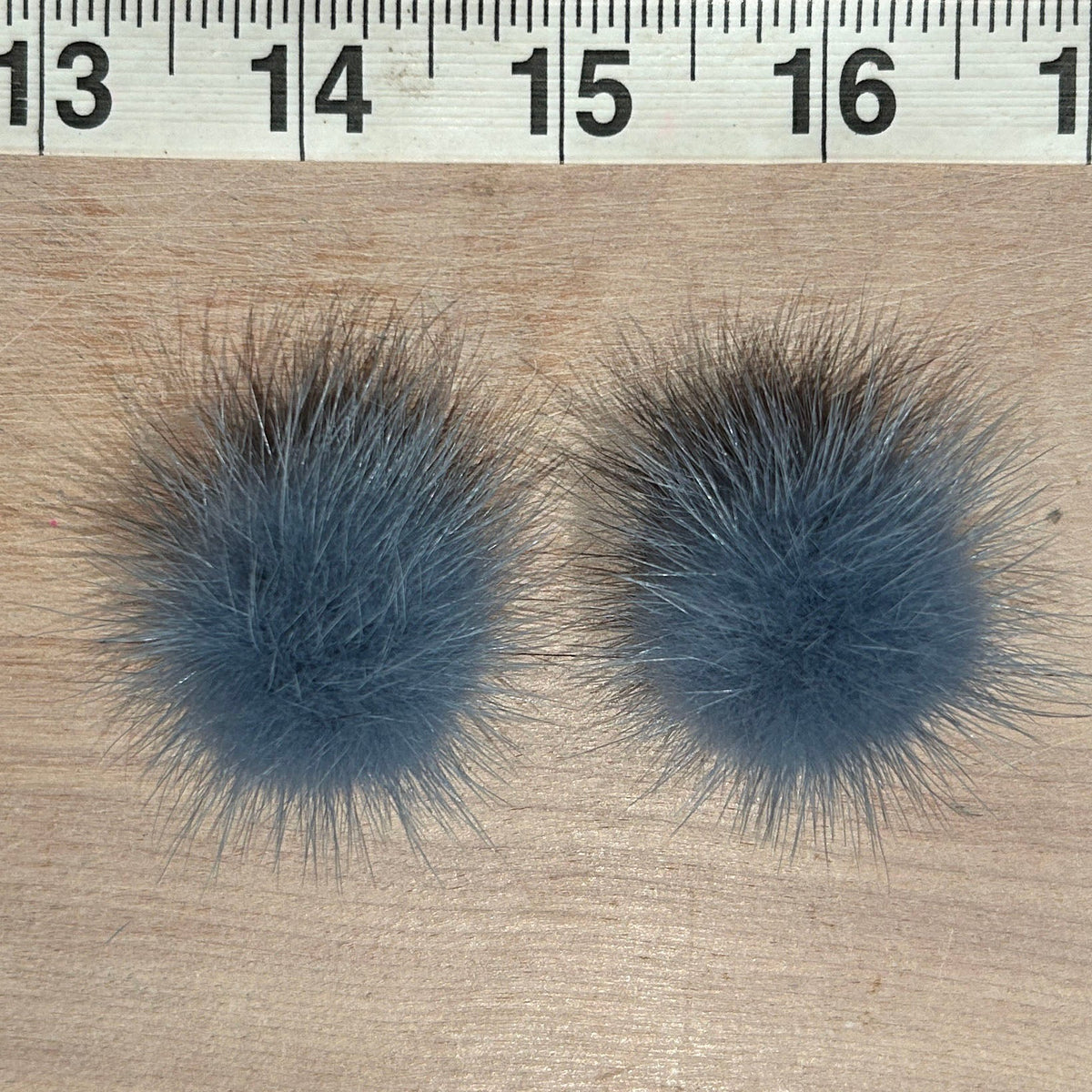 Two round fluffy pom-poms in a dusty slate blue-gray color, shown side-by-side on a light wood surface with a measuring ruler at the top for scale.