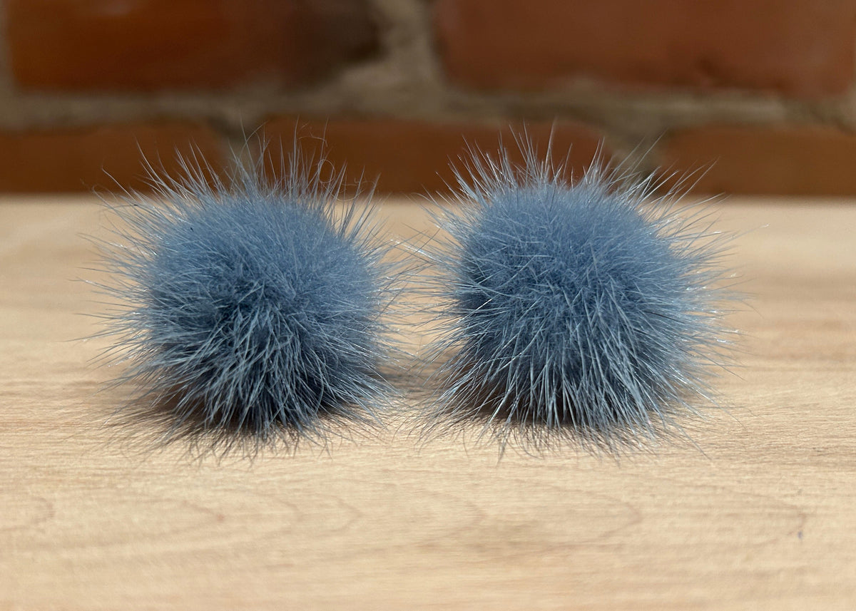 A pair of fluffy blue-gray mink fur pom poms resting on a light wood surface with a blurred brick wall background.