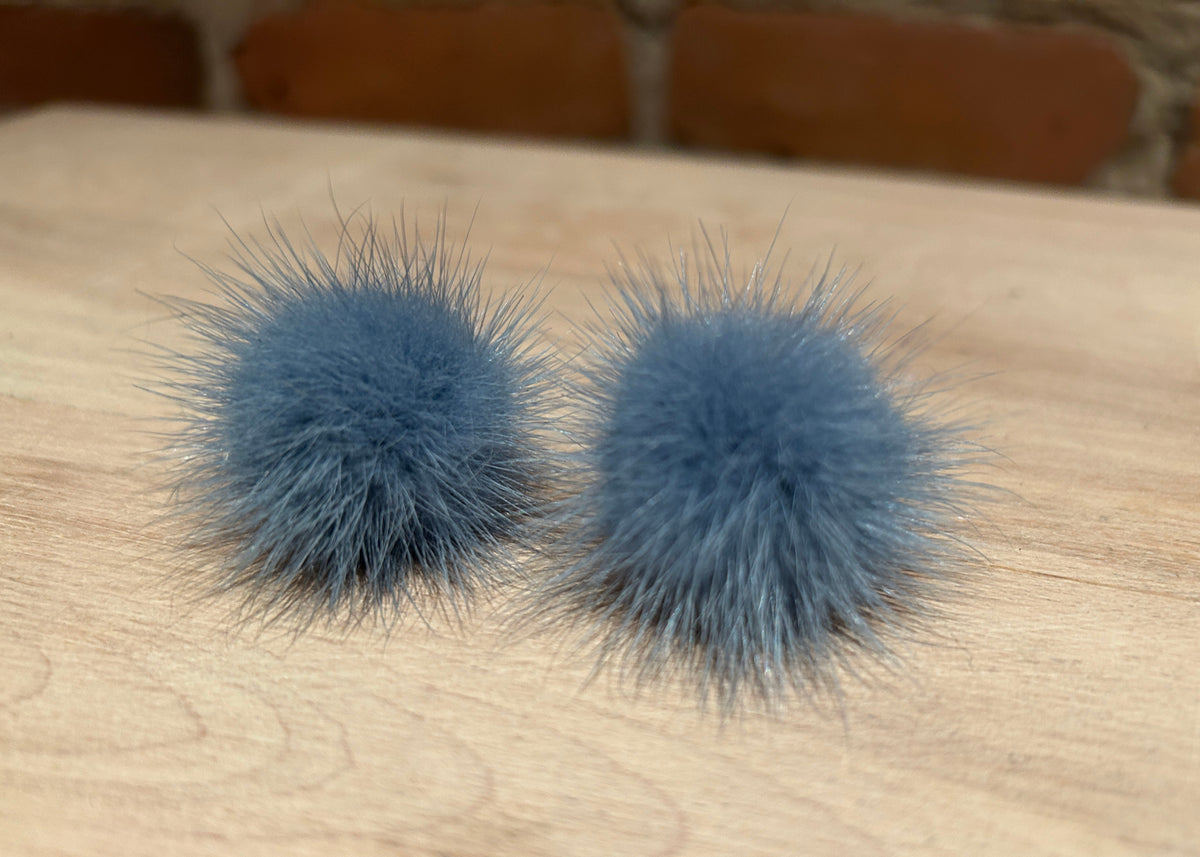 Two fluffy blue-gray mink fur pom poms resting on a light wood tabletop with a blurred brick wall background.