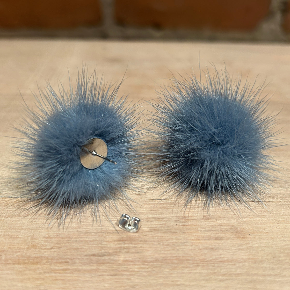 A pair of fluffy pom-pom stud earrings in a dusty slate blue-gray color, displayed on a light wood surface with a brick wall in the background. One earring is shown from the front, while the other is turned to show the metal post and backing.