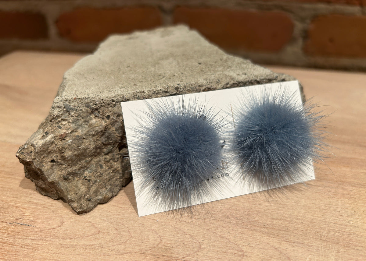 A pair of fluffy pom-pom stud earrings in a dusty slate blue-gray color, displayed on a small white card resting on a wooden surface next to a rough stone, with a blurred brick wall in the background.