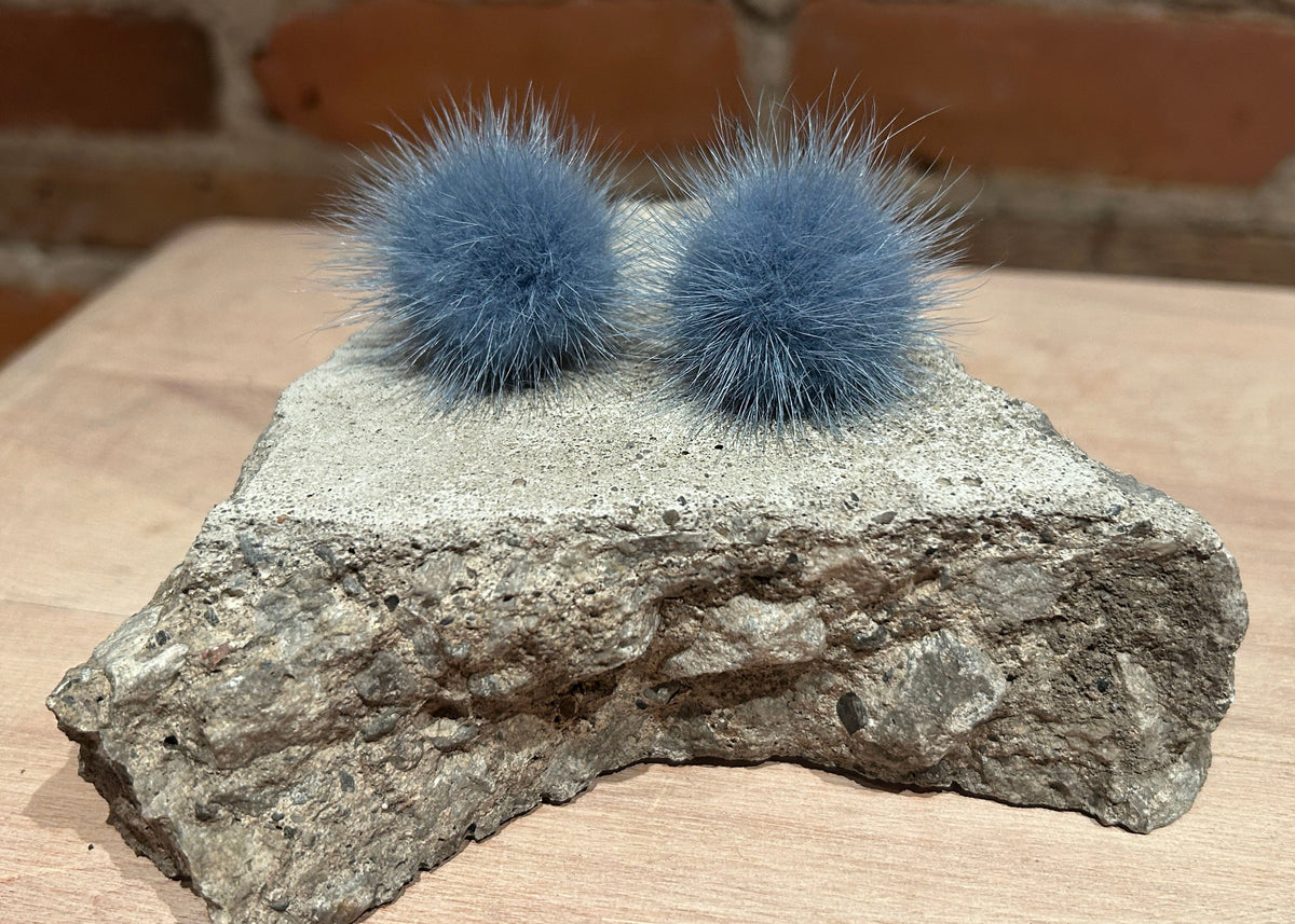 A pair of fluffy blue-gray mink fur pom-poms displayed on top of a rough stone slab placed on a light wood surface, with a blurred brick wall in the background.