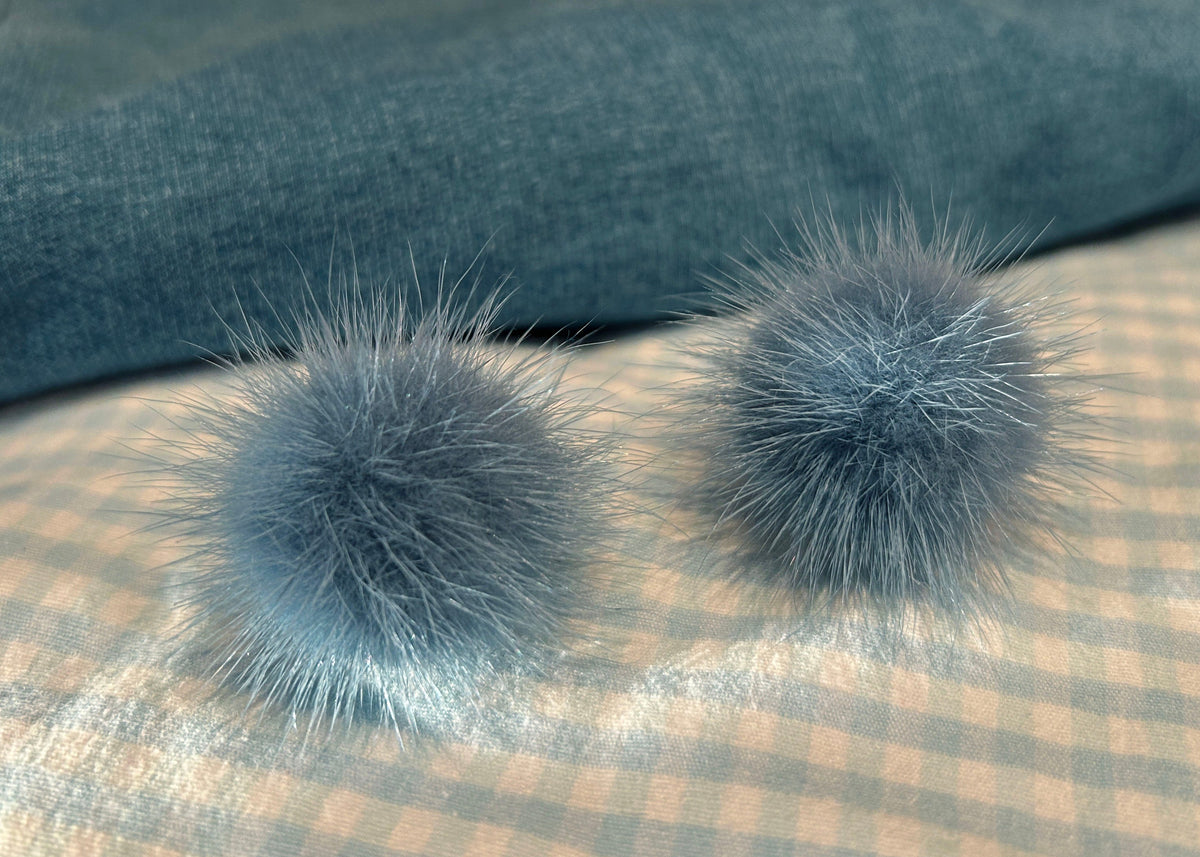 Two fluffy pom-pom earrings in a dusty blue-gray color, resting on a beige and blue checkered fabric surface with a dark blue textured background.