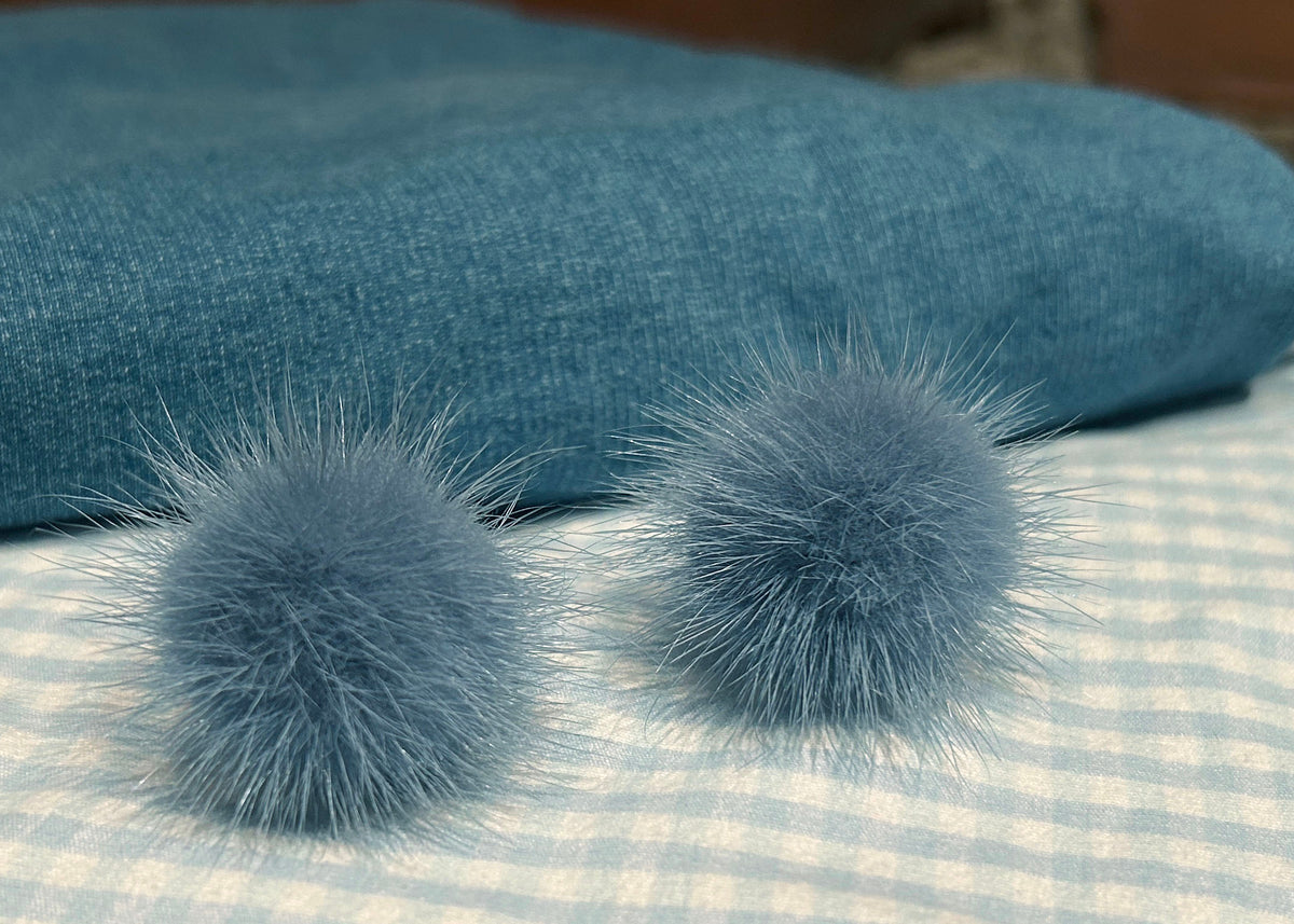 A pair of dusty blue mink fur pom-pom earrings resting on a white and blue gingham fabric surface, with a textured blue fabric accessory in the background.
