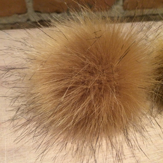A close-up of a fluffy faux-fur pom-pom in a warm tan or light brown color, resting on a light wood surface with a brick wall in the background.
