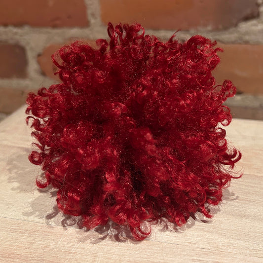 A vibrant deep red curly hat pom pom displayed on a light wood surface with a blurred brick wall background.