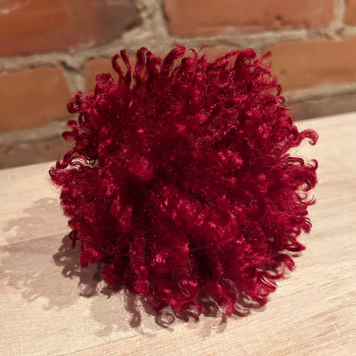 A deep burgundy curly pom-pom accessory for a knit hat, resting on a light wood surface with a brick wall background.