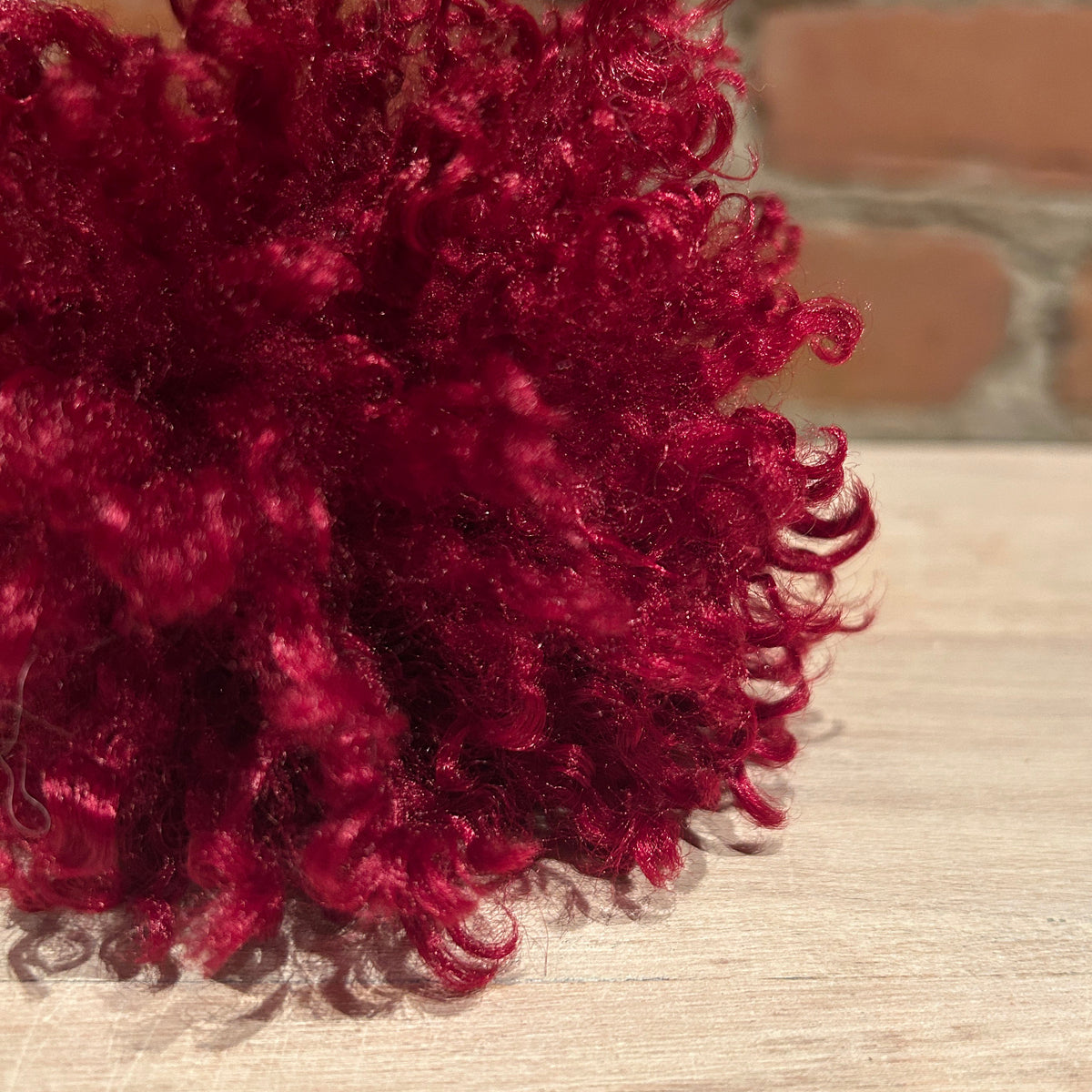 A close-up of a deep burgundy curly hat pom pom resting on a light wood surface with a blurred brick wall background.
