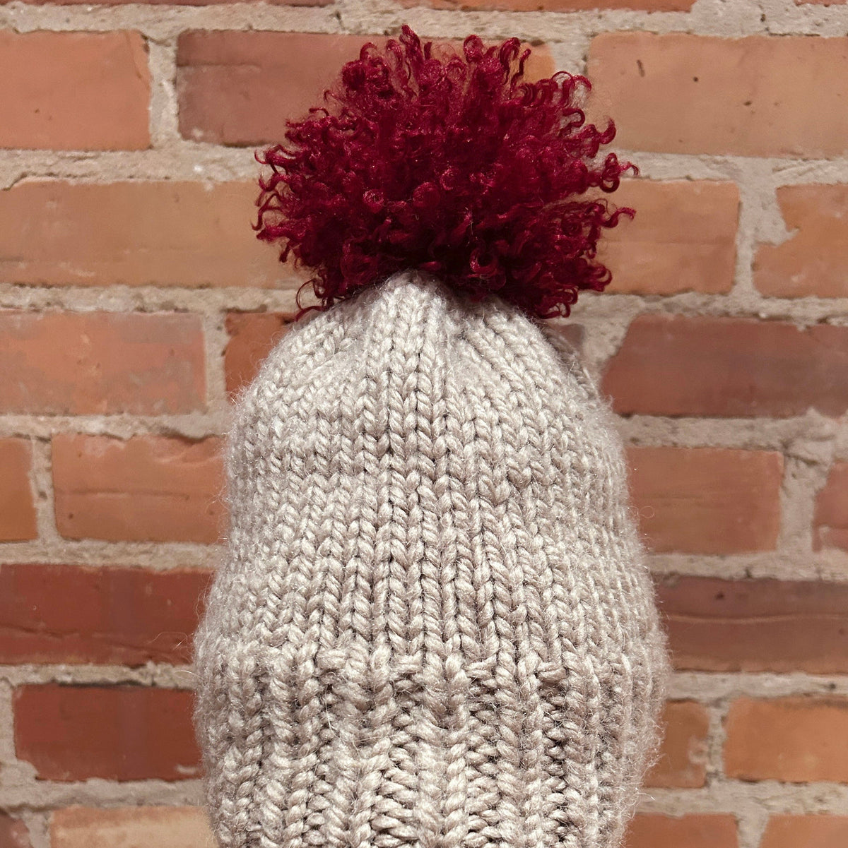 A chunky knit beanie hat in light gray with a dark red pom-pom on top, displayed on a mannequin head against a red brick wall background.