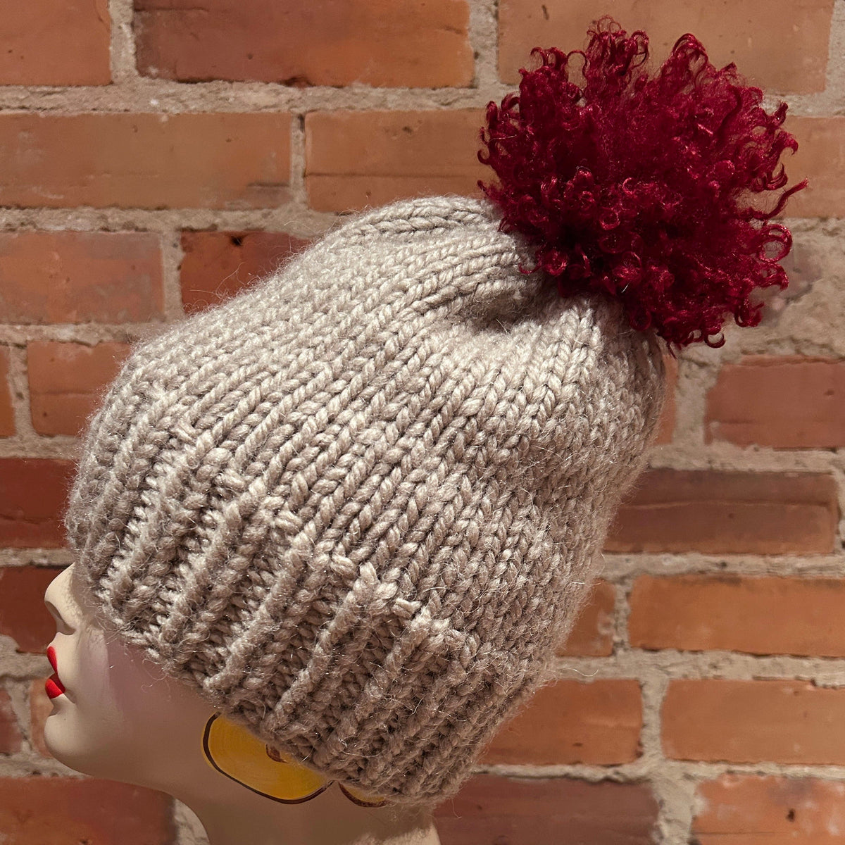 A light gray ribbed knit beanie with a large dark red curly pom-pom, displayed on a mannequin head against a brick wall background.