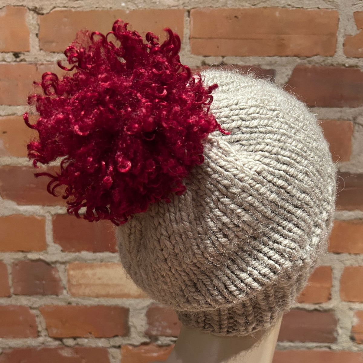 A light beige chunky-knit beanie hat with a large, vibrant red curly pom-pom accessory, displayed on a mannequin head against a brick wall background.