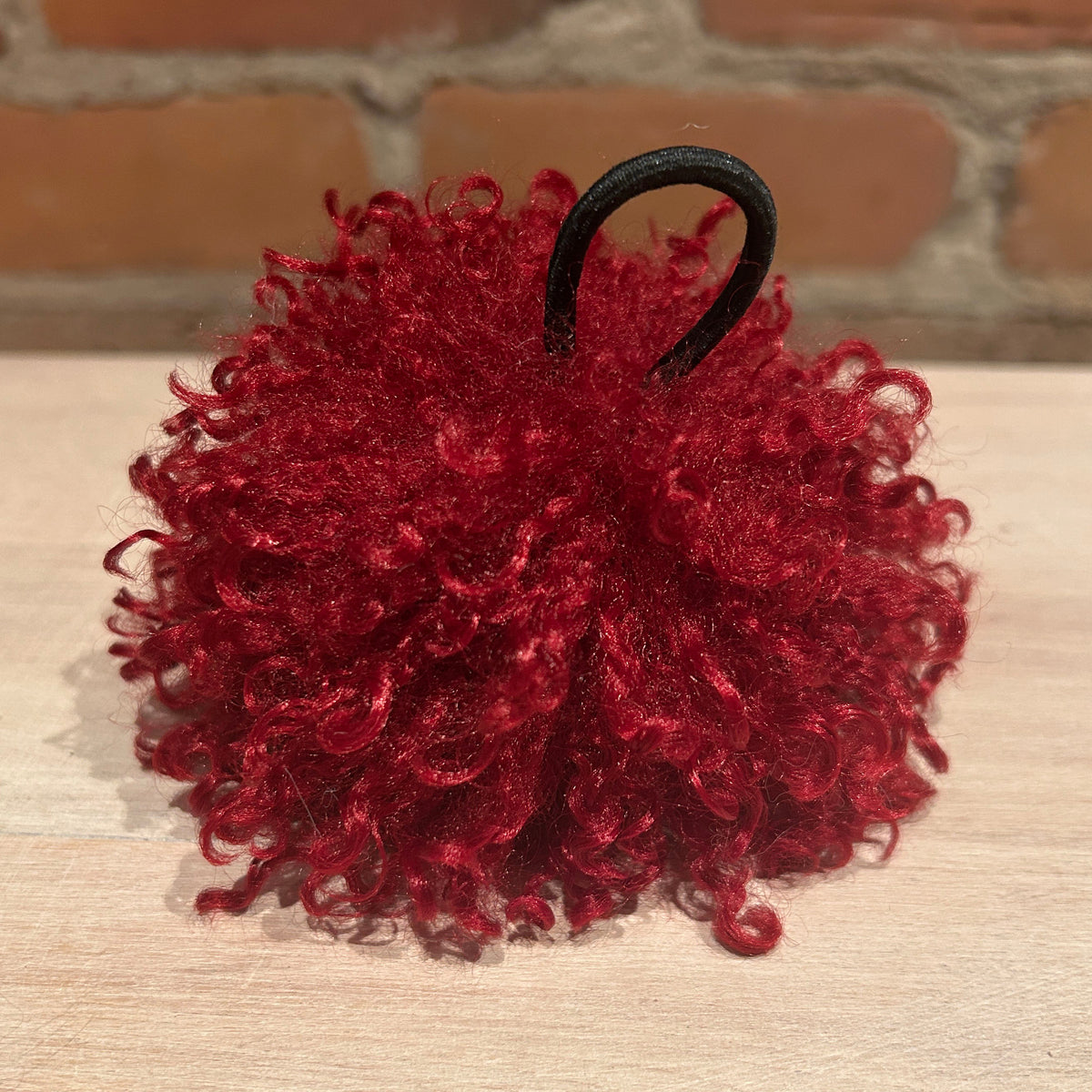 A deep red curly hat pom pom with a black elastic hair tie, displayed on a light wood surface with a brick wall background.