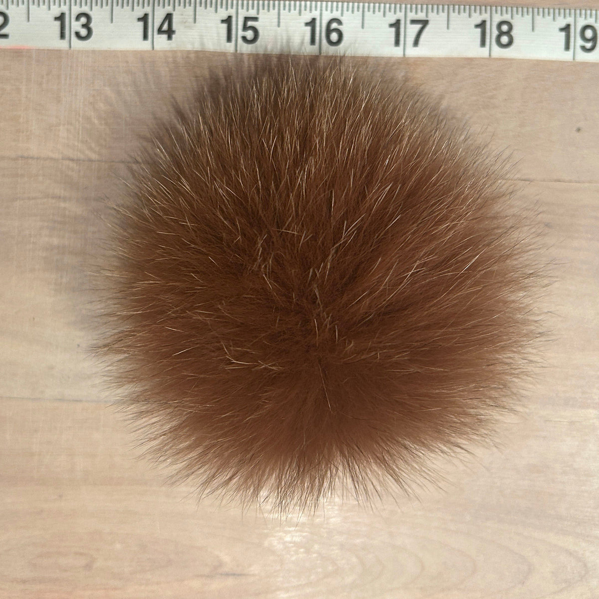 A ginger fox fur pom-pom ball shown on a light wood surface with a measuring tape ruler at the top for scale.