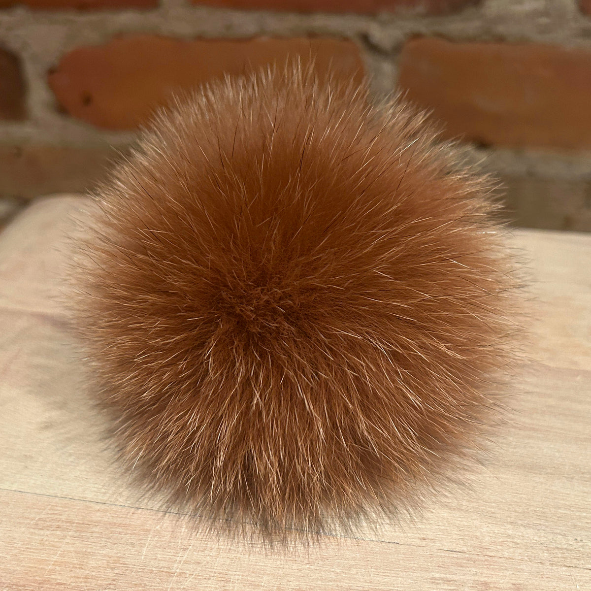 A fluffy, spherical brown fox fur pom-pom resting on a light-colored wooden surface with a blurred brick wall in the background.