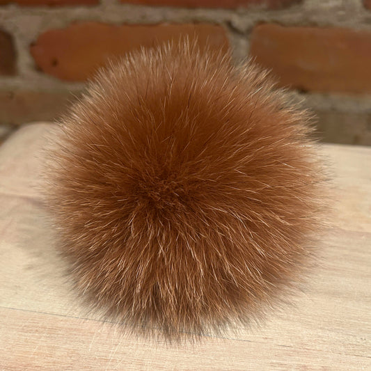 A fluffy, spherical brown fox fur pom-pom resting on a light-colored wooden surface with a blurred brick wall in the background.