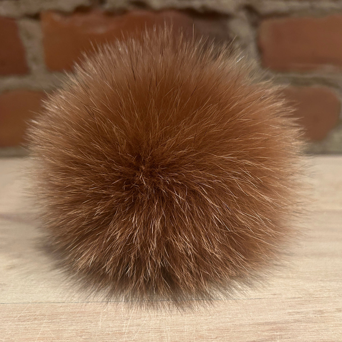 A fluffy, spherical brown fox-fur pom-pom accessory resting on a light wood surface with a blurred brick wall background.
