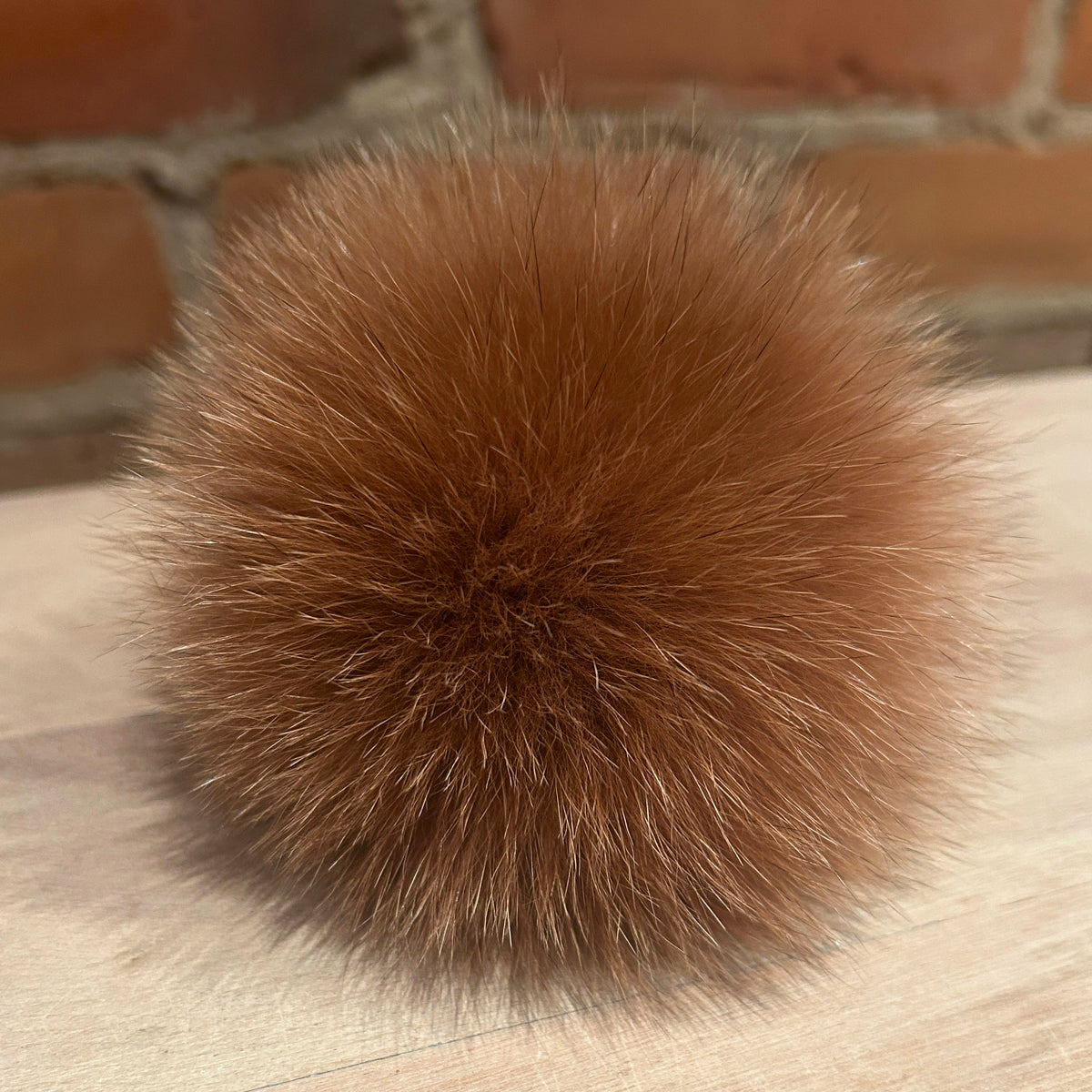 A fluffy, spherical brown fur pom-pom resting on a light-colored wooden surface with a blurred brick wall in the background.