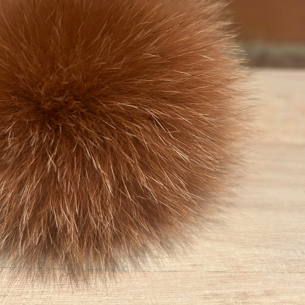 A close-up of a fluffy brown fox fur pom-pom resting on a light wood surface with a blurred brick wall in the background.