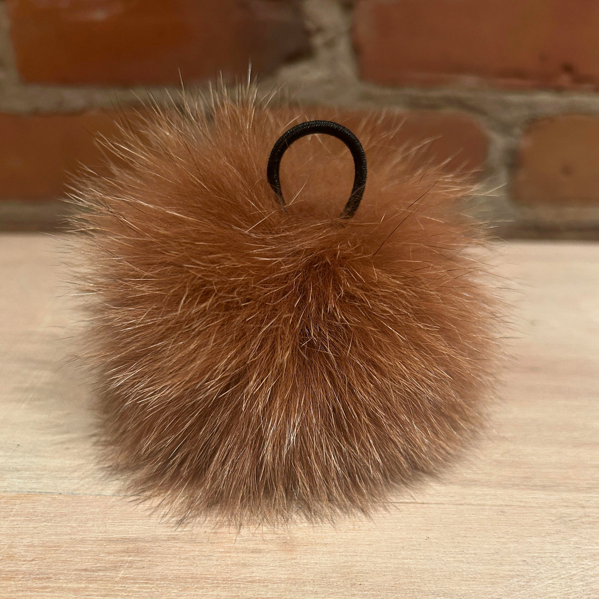 A ginger fox fur pom-pom keychain or bag charm with a black elastic loop, resting on a light wood surface with a brick wall background.